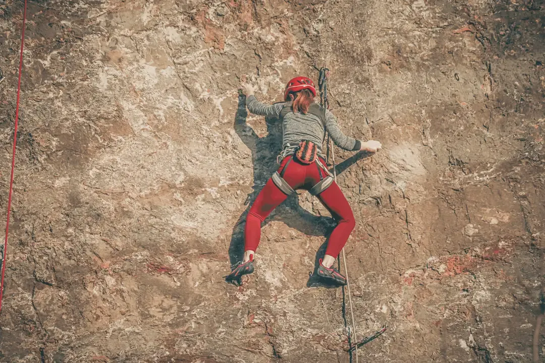 Escaladora con casco rojo, vista desde abajo y de espaldas, escalando de primera una vía deportiva en una pared de roca al aire libre