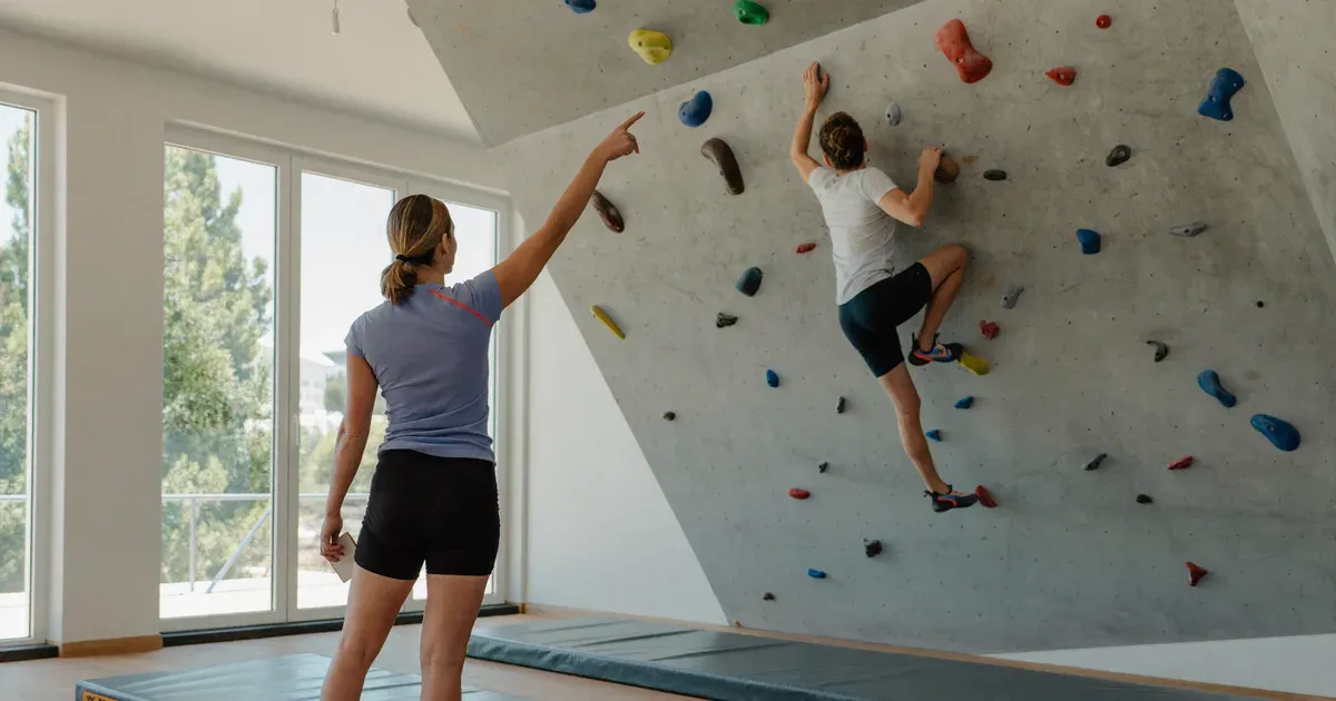 Entrenadora de pie sobre la colchoneta señalando una presa mientras una escaladora avanza en el muro de bloque de un rocódromo luminoso