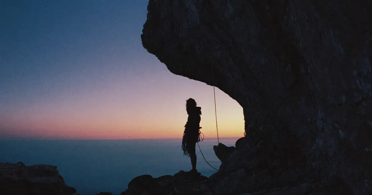 Silueta al pie de una pared rocosa al atardecer, con la cuerda preparada y el mar al fondo