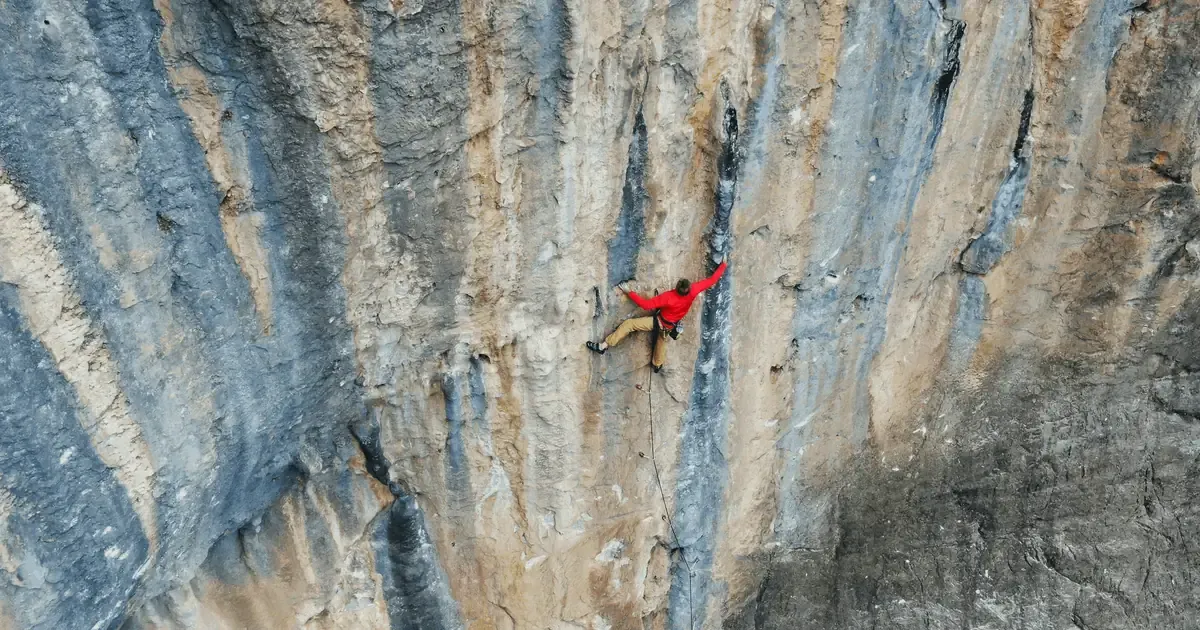 Vista aérea de un escalador con sudadera roja en mitad de una vía deportiva, sobre una pared de caliza gris y ocre con chorreras