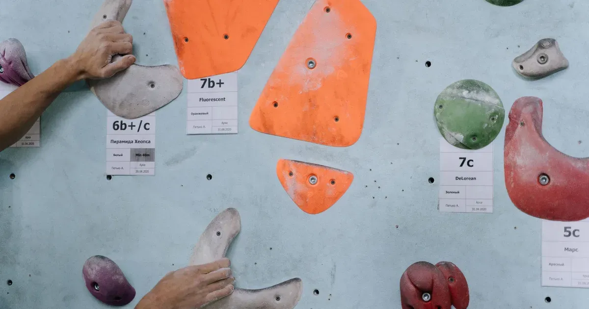 Dos manos con magnesio agarrando presas en un muro de bloque, con las etiquetas de grado de las vías -6b+/c, 7b+, 7c- pegadas en la pared junto a cada presa